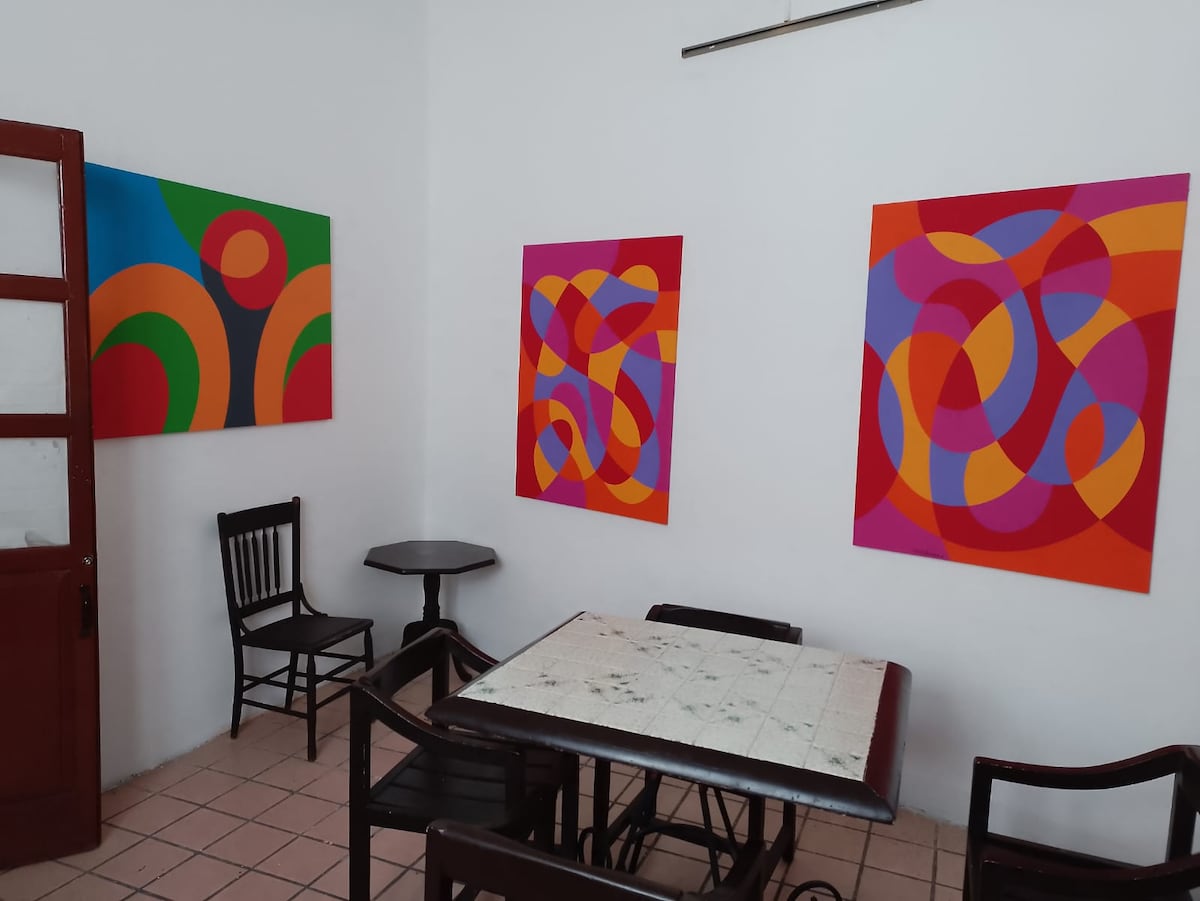 A bright dining area features a square table surrounded by black wooden chairs. Three vibrant abstract paintings adorn the white walls, showcasing bold colors and shapes. The tiled floor adds a simple yet complementary element to the space.