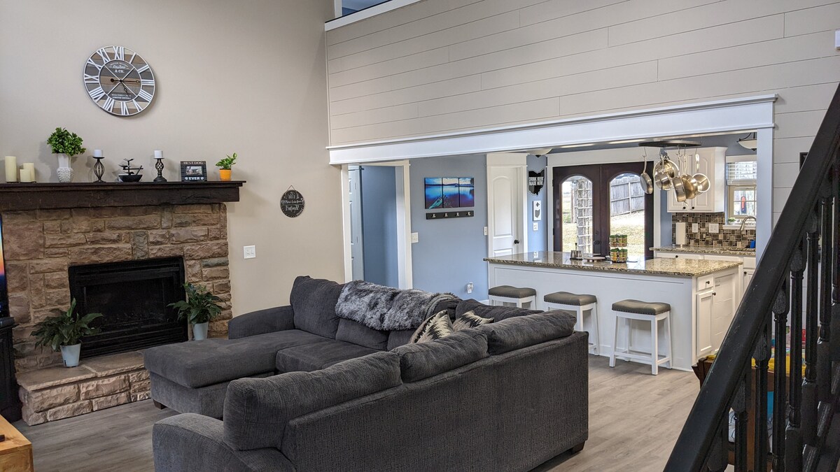 A spacious living room features a large sectional sofa that comfortably accommodates two people. The area is complemented by a stone fireplace, and light streams in from the adjacent open-concept kitchen fitted with modern bar stools. A wall clock adds a decorative touch.