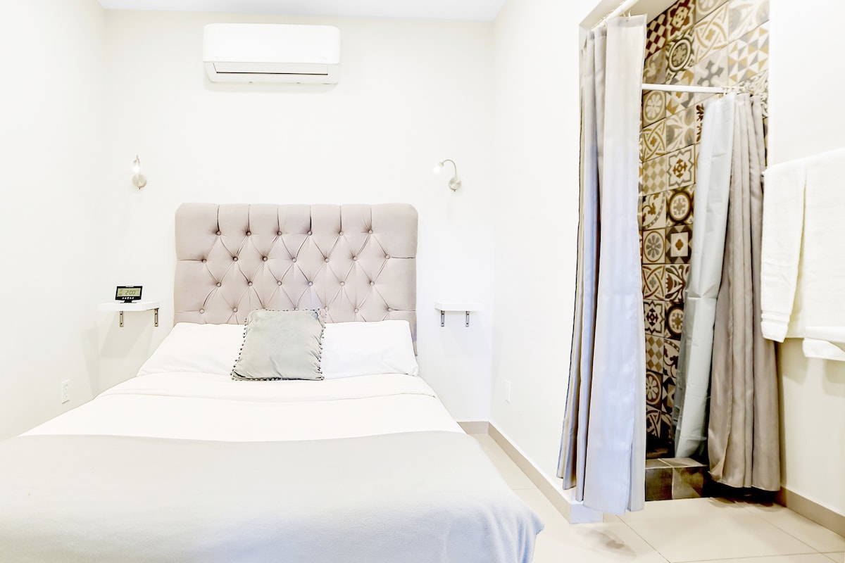 A well-appointed sleeping area features a plush bed with an elegantly tufted headboard. A private bathroom is visible through a fabric curtain, showcasing decorative tiles. Natural light filters through the space, complemented by a wall-mounted air conditioning unit.