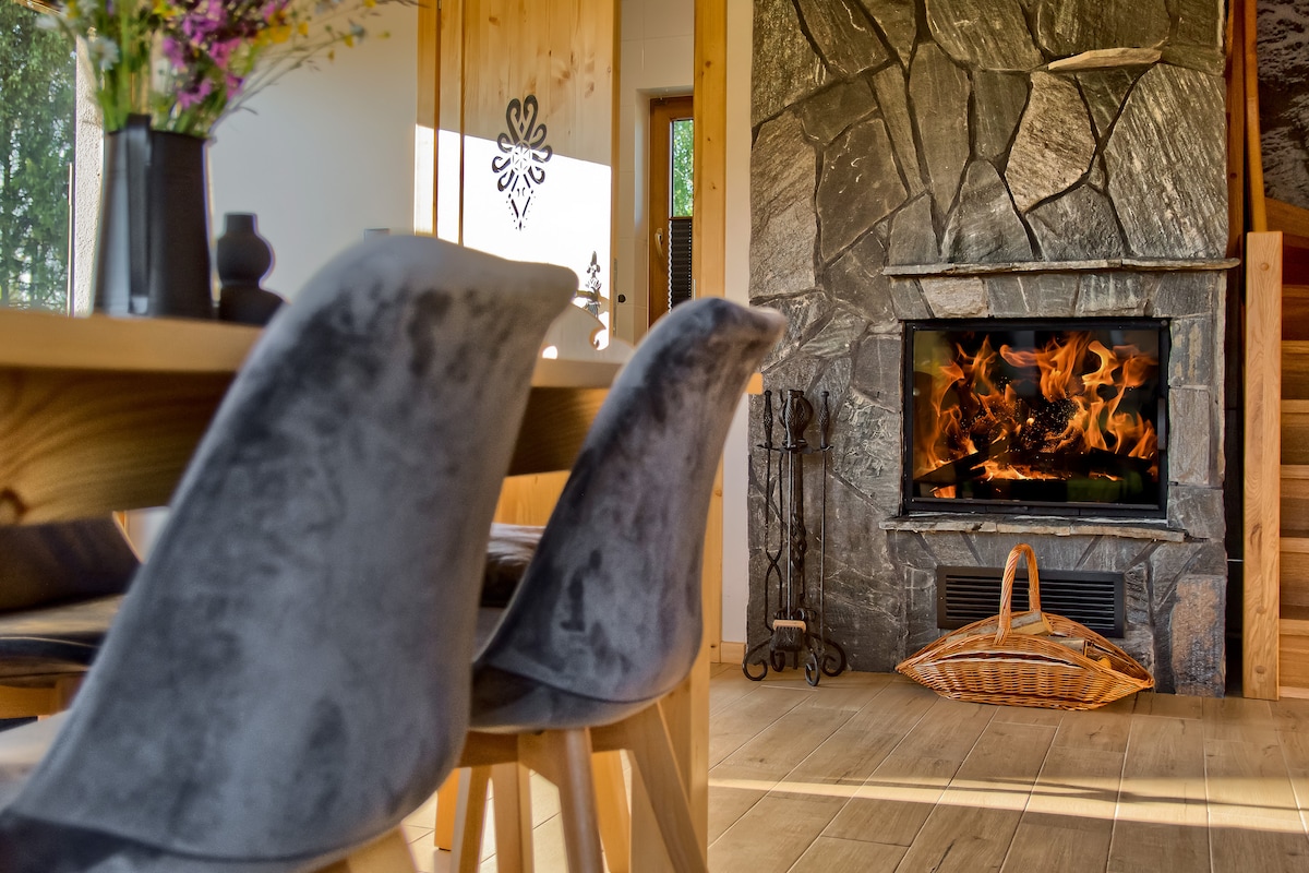 A warm living area is featured with a stone fireplace, flames flickering within. Grey chairs surround a wooden dining table, which is complemented by a decorative arrangement of flowers. Natural light filters through nearby windows, adding a soft glow to the interior.