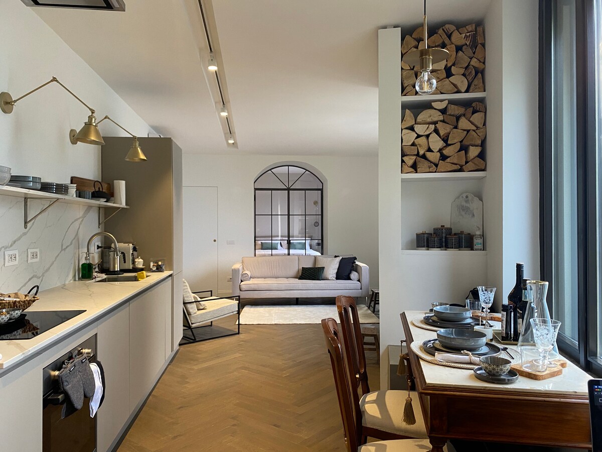 A modern kitchen area is shown, featuring a long countertop with cooking utensils and a stylish seating space. A cozy living area with a sofa and a large window offers a view of the outdoors. Decorative wooden logs are neatly arranged in a shelf.