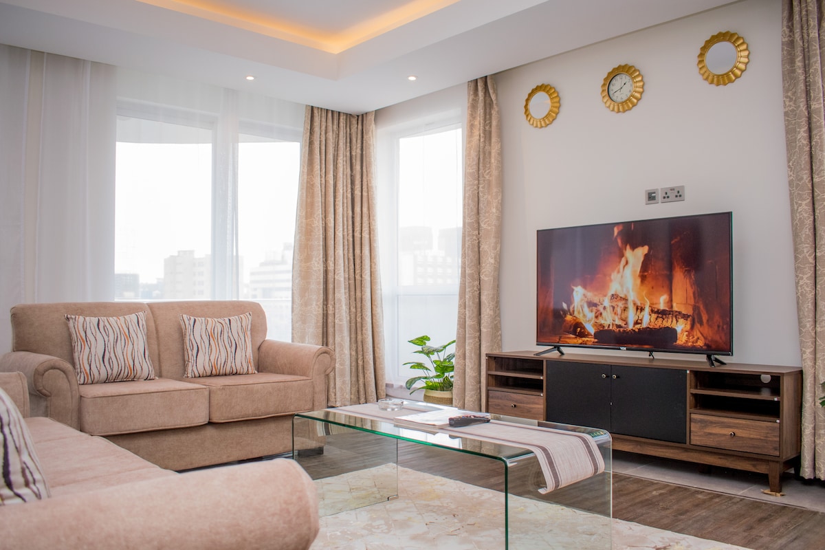 An inviting living area features a comfortable sofa adorned with patterned cushions, paired with a glass coffee table. A modern television is positioned in front of the sofa, complemented by an elegant decor of golden wall clocks. Large windows allow ample natural light.