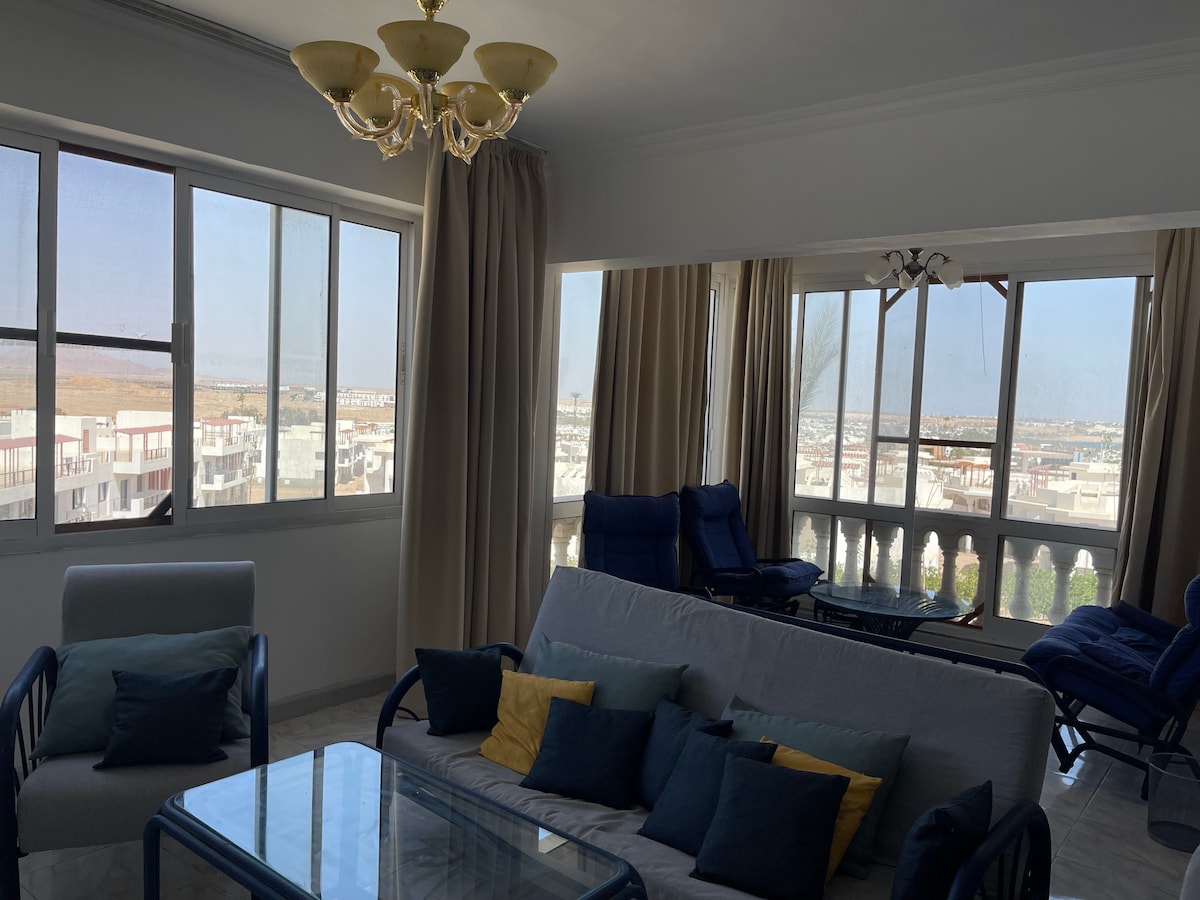 A bright living area features large windows that allow natural light to fill the space. Comfortable seating with decorative cushions is arranged around a glass coffee table. Soft drapes frame the windows, enhancing the panoramic views of the surrounding area.