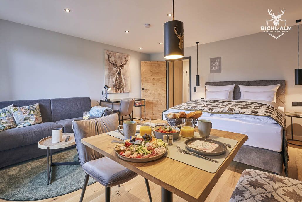 A well-furnished one-room apartment features a cozy living space with a comfortable sofa and dining area. A spacious bed is prominently displayed, complemented by a beautiful breakfast spread on the table. Natural light fills the room, enhancing its inviting atmosphere.