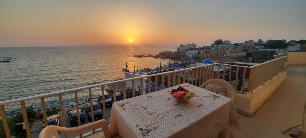 A balcony offers a view of the setting sun over the ocean, casting warm colors across the sky. A table with a tablecloth is set for two and displays a bowl of fresh fruit, enhancing the coastal experience.