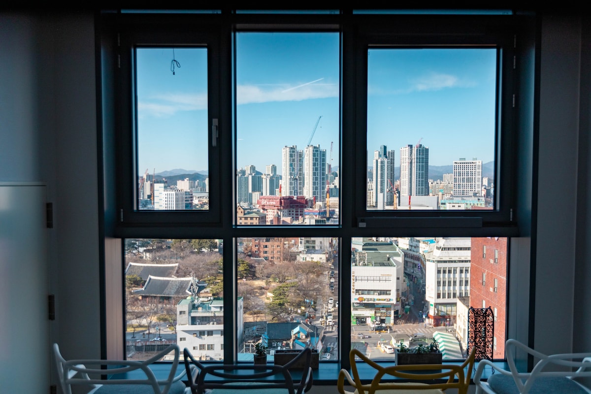 A large window provides a view of the city skyline, featuring tall buildings and distant mountains. The scene is illuminated by natural light, creating a bright and spacious atmosphere in the interior of the room.