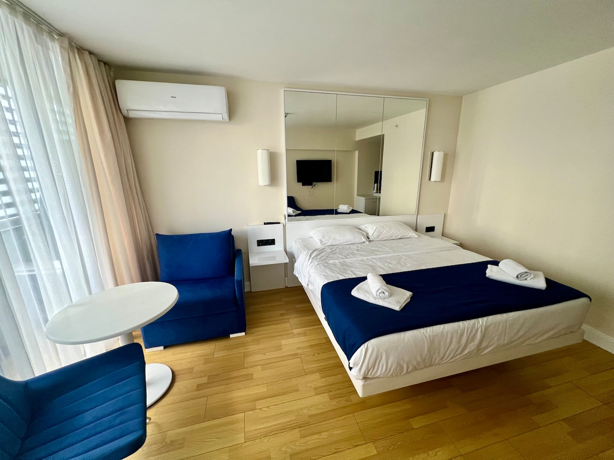A bright studio apartment features a spacious bed covered with white linens and a navy blue throw. A matching blue armchair is situated next to a round white table. A large mirror reflects the natural light and enhances the openness of the space.