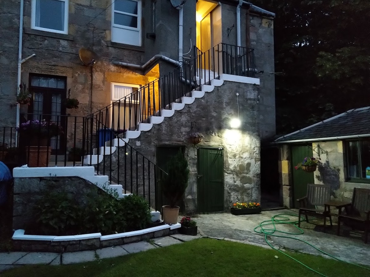 An exterior view of the property reveals a stone building with illuminated stairs leading to the entrance. Lush greenery surrounds the area, complemented by flower planters and a well-lit outhouse. A patio with seating offers a serene space for relaxation.