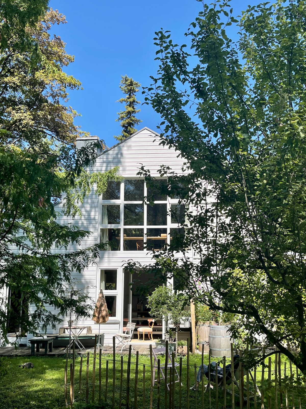 A modern house with large, tall windows is framed by lush green trees. The outdoor space features seating arrangements on a manicured lawn, creating a peaceful setting that invites relaxation. Natural light brightens the exterior, enhancing the connection to the surrounding nature.