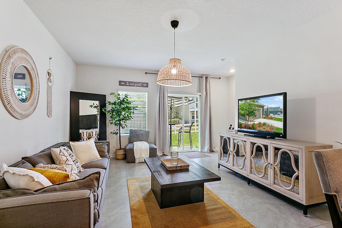 A spacious living area features comfortable seating arranged around a central coffee table. A large television is positioned on a stylish cabinet, while a decorative light fixture adds interest above. Natural light filters through sliding glass doors, leading to an outdoor space, complemented by greenery.