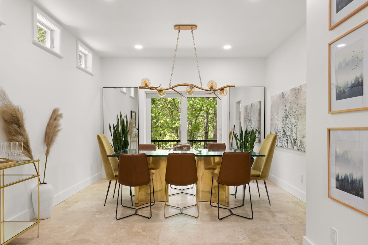 A modern dining area is highlighted by a large glass table surrounded by six brown and gold chairs. Two vibrant green plants are positioned on either side of the table. Large windows allow natural light to fill the space, providing views of the outdoors.