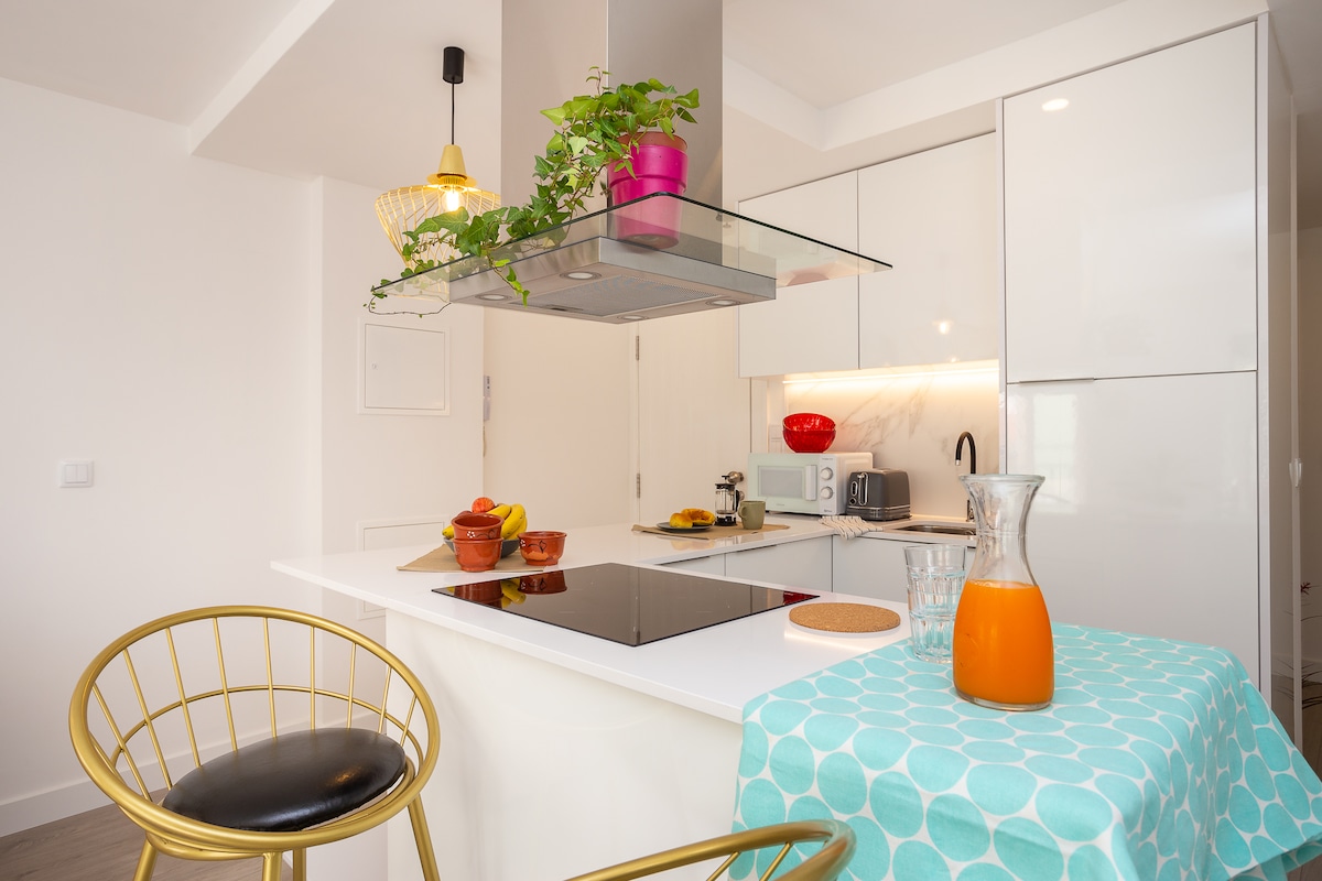 A modern kitchen is presented with sleek white cabinetry and a stylish island featuring a stovetop. Gold-accented stools provide seating, while a table with a turquoise cloth displays a pitcher of orange juice and a bowl of fruit. Greenery is added with a potted plant above the island.
