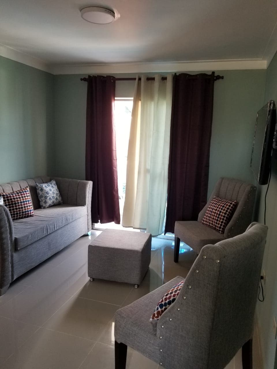 A cozy seating area is furnished with a gray sofa and two matching chairs, featuring decorative pillows. A small gray ottoman is positioned in the center. Natural light filters through a window, adorned with sheer and dark curtains, illuminating the light green walls.