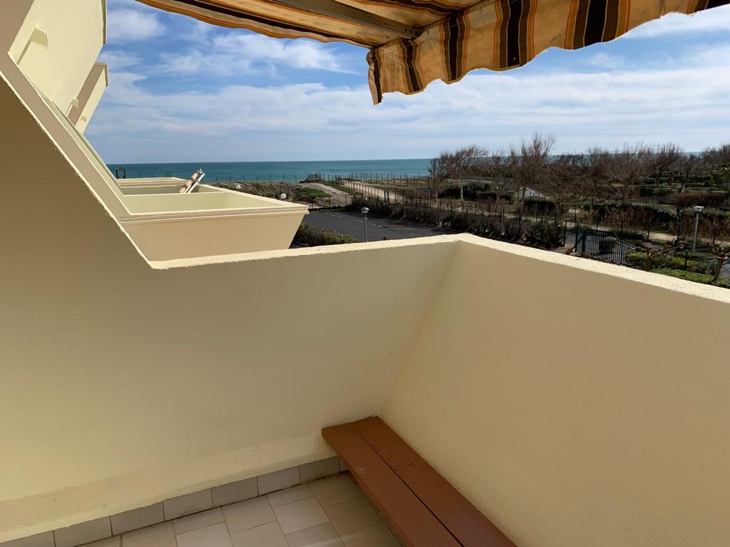 A terrace with a bench offers a clear view of the sea, framed by a striped awning. The foreground showcases the terrace's tile flooring, while the background reveals the horizon meeting the ocean. Vegetation and pathways can be seen along the beachfront.