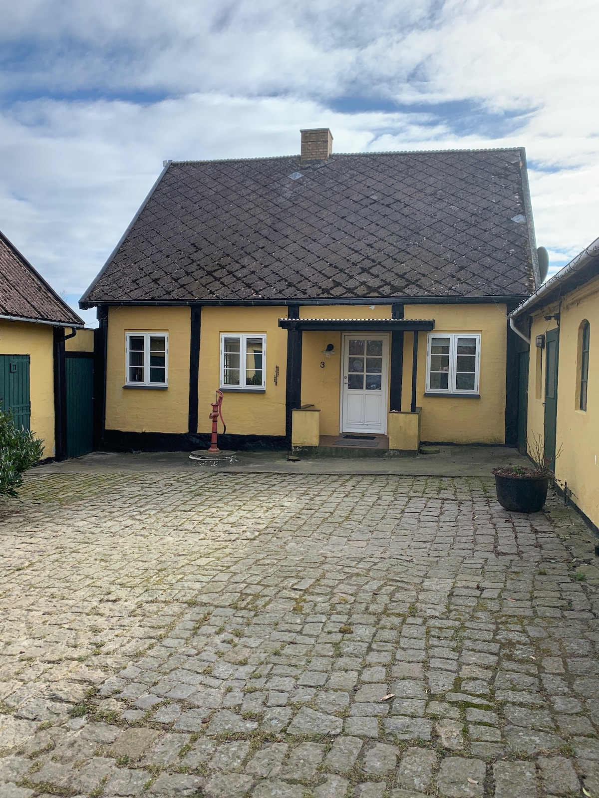 A quaint yellow house features a traditional sloped roof and a front entryway with white trim. Several small windows are visible, allowing natural light to enhance the entrance. The surrounding cobblestone courtyard is complemented by greenery in pots and offers a peaceful atmosphere.