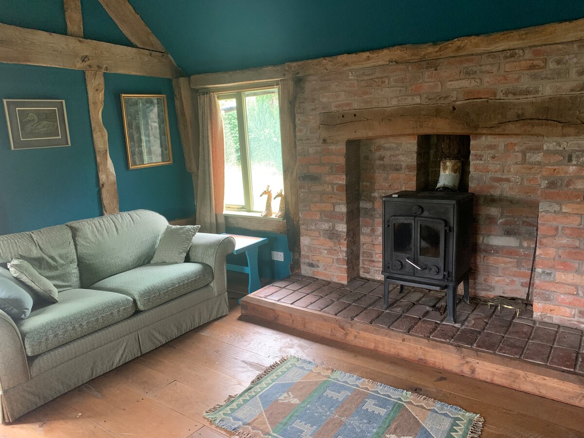 A cozy lounge area is highlighted by a brick fireplace with a wood-burning stove. A fabric sofa with decorative cushions faces the fireplace, while a small rug adds texture to the wooden floor. Natural light enters through a window dressed with light curtains.