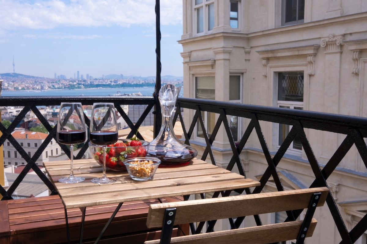 A private balcony features a wooden table set with two glasses of red wine, a decanter, strawberries, and nuts. The surrounding view includes the Bosphorus and the city skyline, framed by the elegant design of the building's architecture.