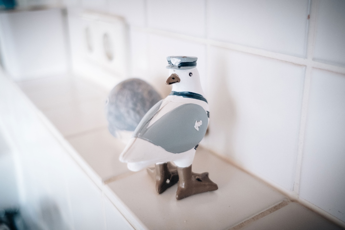 A decorative ceramic bird figurine is placed on a white tiled surface, featuring a gray and white color scheme with a small blue hat. A spherical object can be seen nearby, also on the tiled surface.