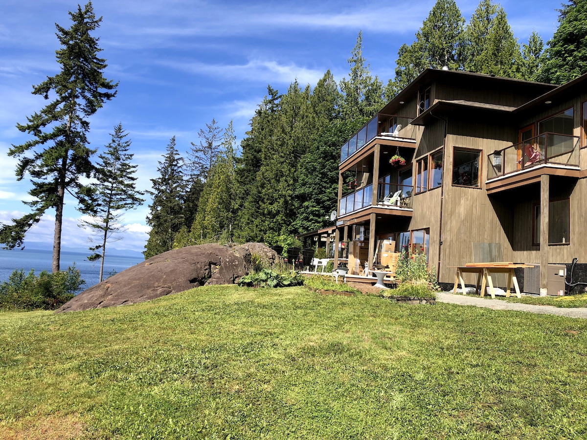 The image showcases a multi-level building set against a backdrop of lush green trees and ocean views. A large smooth rock and well-kept lawn are visible in the foreground, while spacious decks and patio furniture are seen on the exterior of the building.