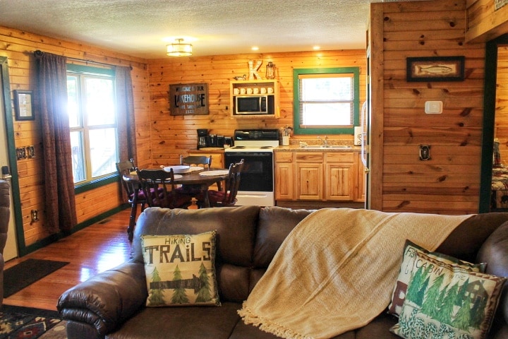 A cozy living area features a brown leather sofa draped with a knitted blanket. Natural wood walls create a warm atmosphere, while a dining table is situated nearby. The kitchen area is equipped with appliances and wood cabinetry, with natural light entering through multiple windows.