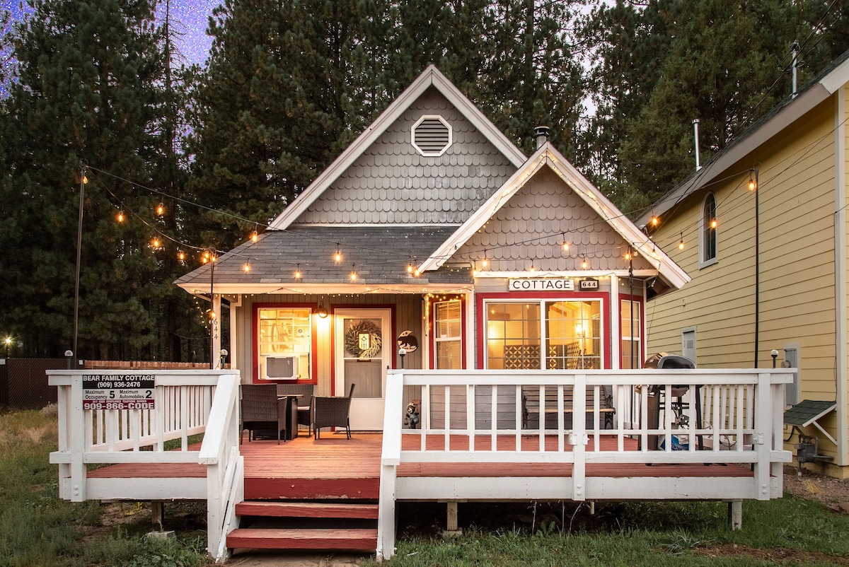 A charming single-story cabin is seen featuring a welcoming front porch adorned with string lights. The rustic wooden deck and light grey shingle roof contribute to the cozy aesthetic. Nearby, tall pine trees provide a natural backdrop, enhancing the cabin's inviting appeal.
