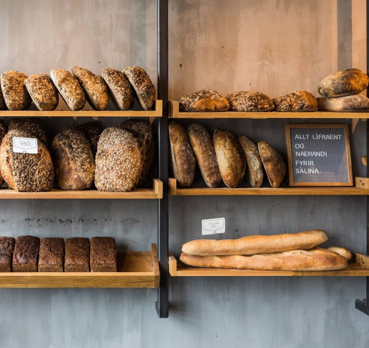 Wooden shelves display an assortment of freshly baked breads, including round loaves topped with seeds and baguette-style bread. A small blackboard sign notes the offerings in Icelandic, adding a local touch. The backdrop features a textured grey wall, enhancing the rustic ambiance.