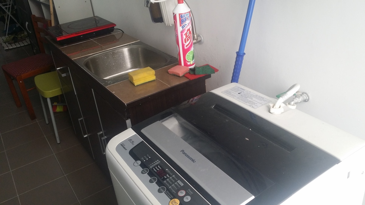 A compact kitchen area features a washing machine and a small sink, complemented by a countertop. Cleaning supplies are visible alongside a dish sponge. A few colorful chairs are positioned nearby, enhancing the practicality of the space.