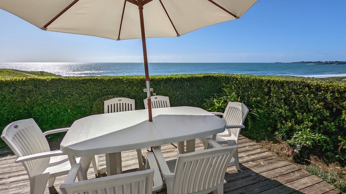 A wooden terrace features a large white table surrounded by six white chairs, all under a large umbrella. The backdrop showcases a clear view of the sea and the horizon, with lush greenery along the edges enhancing the outdoor dining space.