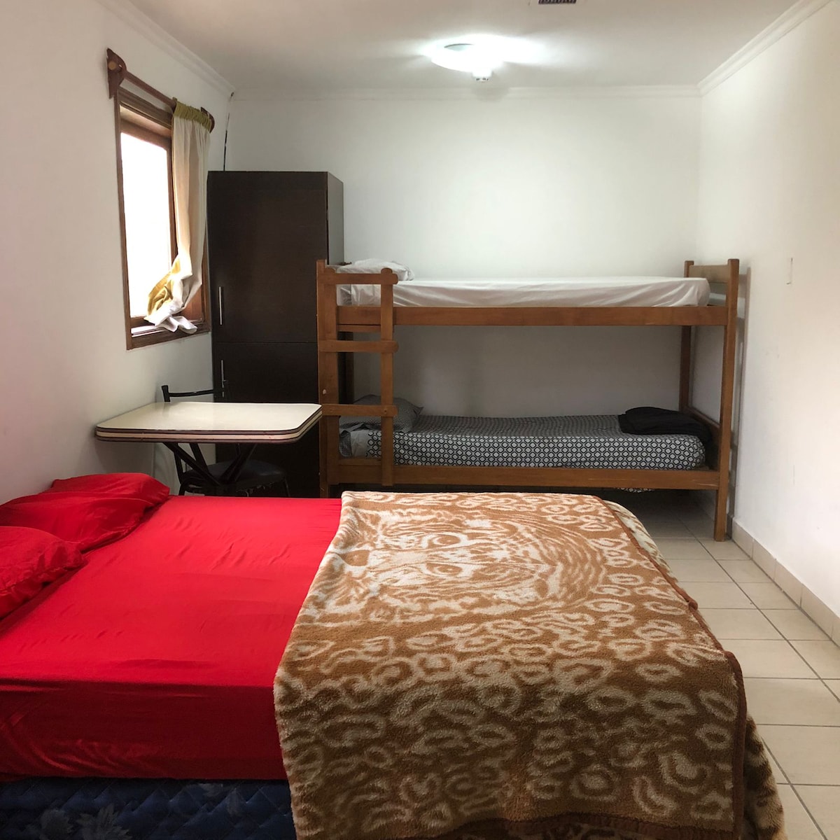 The sleeping area includes a spacious bed with red bedding and a cozy blanket draped over it. A bunk bed is positioned in the corner, complemented by a small table and chair nearby. Natural light enters through the window, brightening the neutral walls.