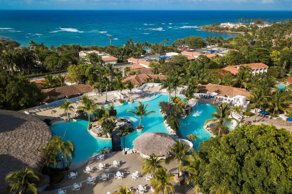 An aerial view of a vibrant beachfront resort, showcasing a large lagoon-style pool surrounded by lush greenery and palm trees. The clear blue water reflects the sky, while pathways weave through the area, leading to shaded loungers and sandy beaches.