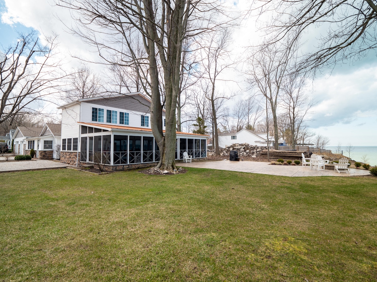 The exterior of a spacious two-story house is shown, featuring a large screened-in porch that overlooks a well-maintained lawn. The property is bordered by tall trees and offers access to Lake Erie, with seating provided on a stone patio nearby.