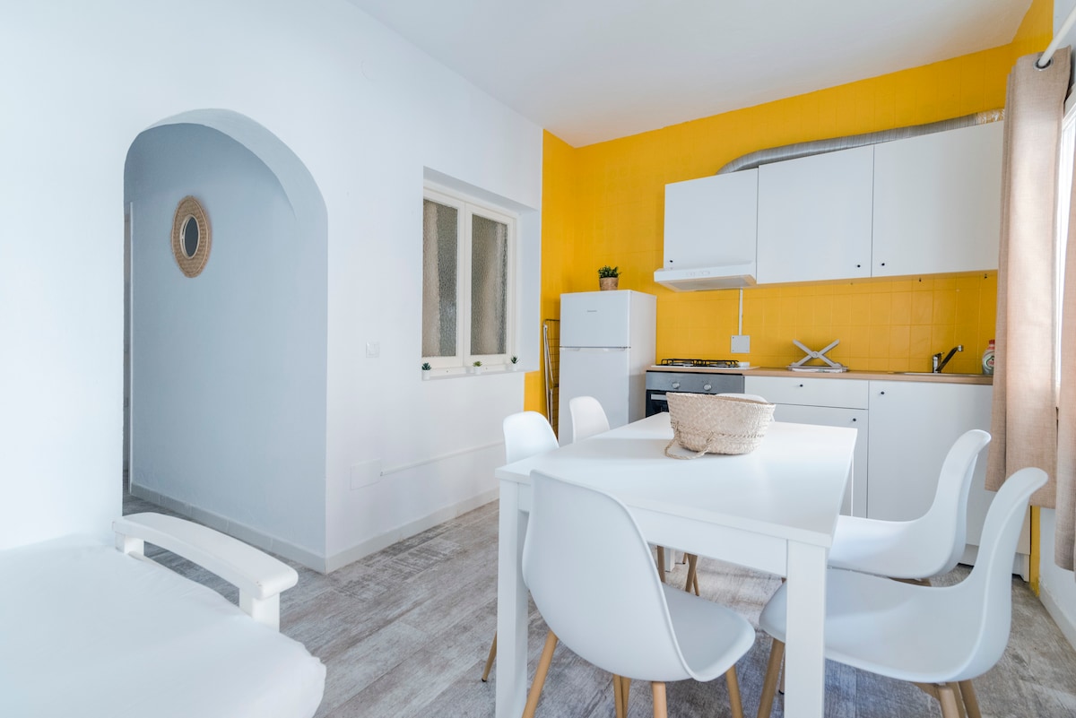 An open kitchen and dining area features a white table surrounded by five chairs. Bright yellow walls contrast with the white cabinetry. The kitchen appliances, including a refrigerator, are neatly arranged, while natural light enters through a window, enhancing the cheerful ambiance.