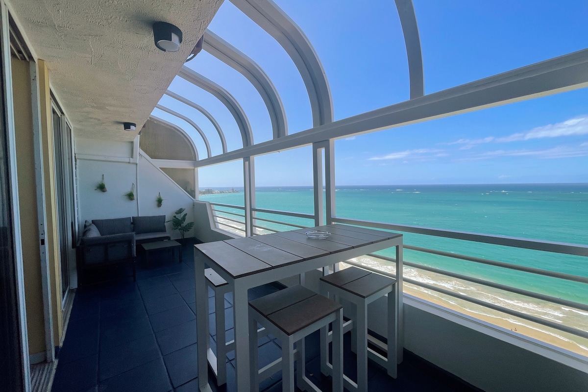 A spacious balcony offers stunning ocean views through large glass panels. A dining table with high stools stands in the center, surrounded by comfortable seating. The serene blue of the water contrasts with the beach, creating a calming atmosphere.