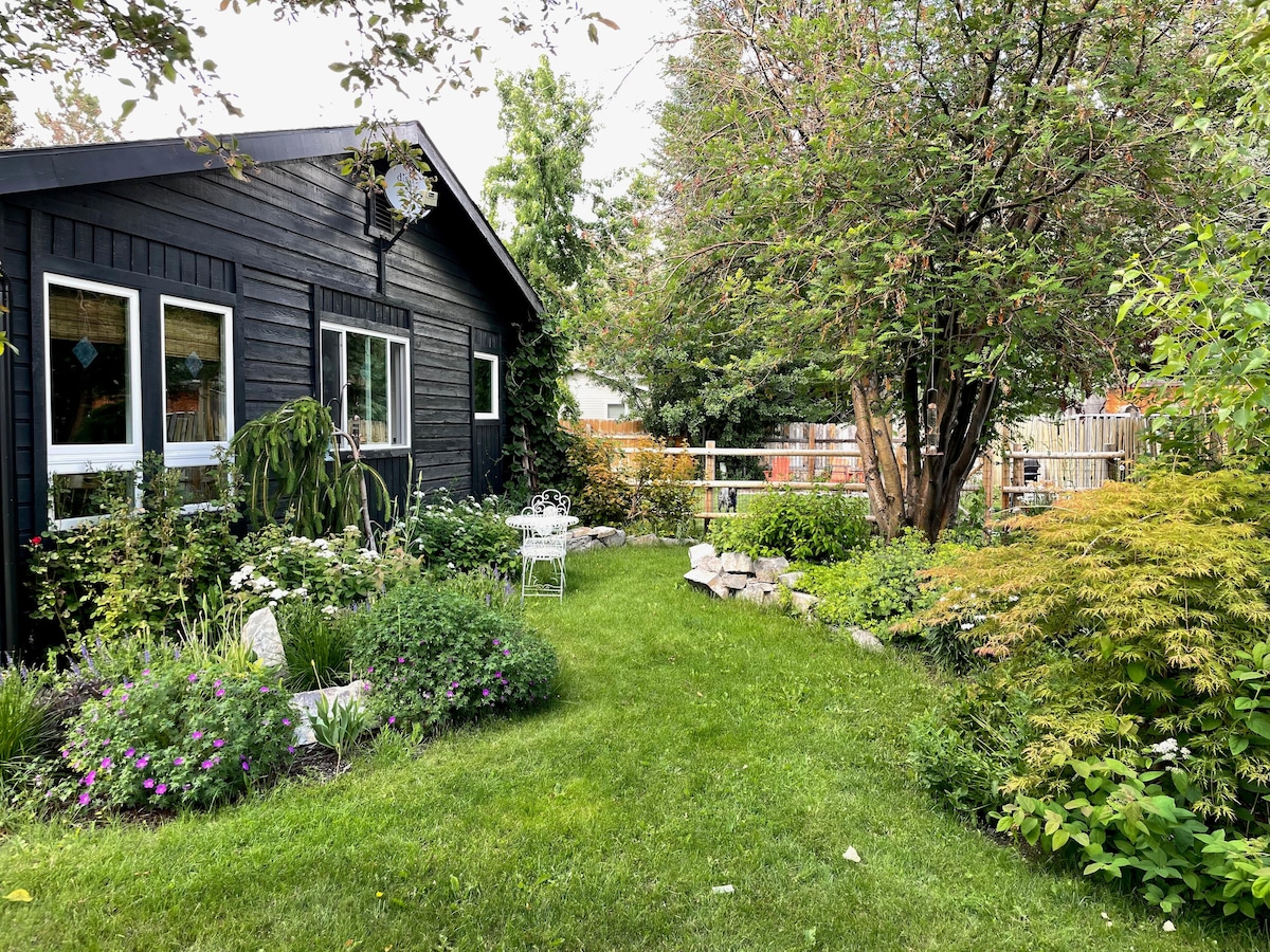 A lush garden surrounds a dark wooden house, featuring vibrant flowers and well-maintained greenery. A few white outdoor chairs are visible, set among the plants. The space offers a tranquil atmosphere with a pathway meandering through the colorful foliage.