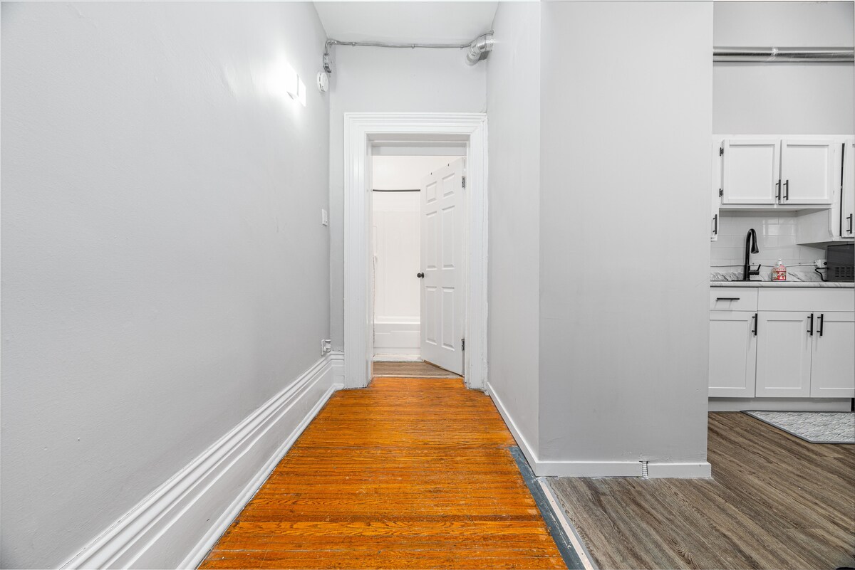 A long hallway features warm wooden flooring leading to a light-colored doorway at the back. Neutral gray walls enhance the spaciousness, while an open door on the right leads to a kitchen area with modern cabinetry and fixtures.