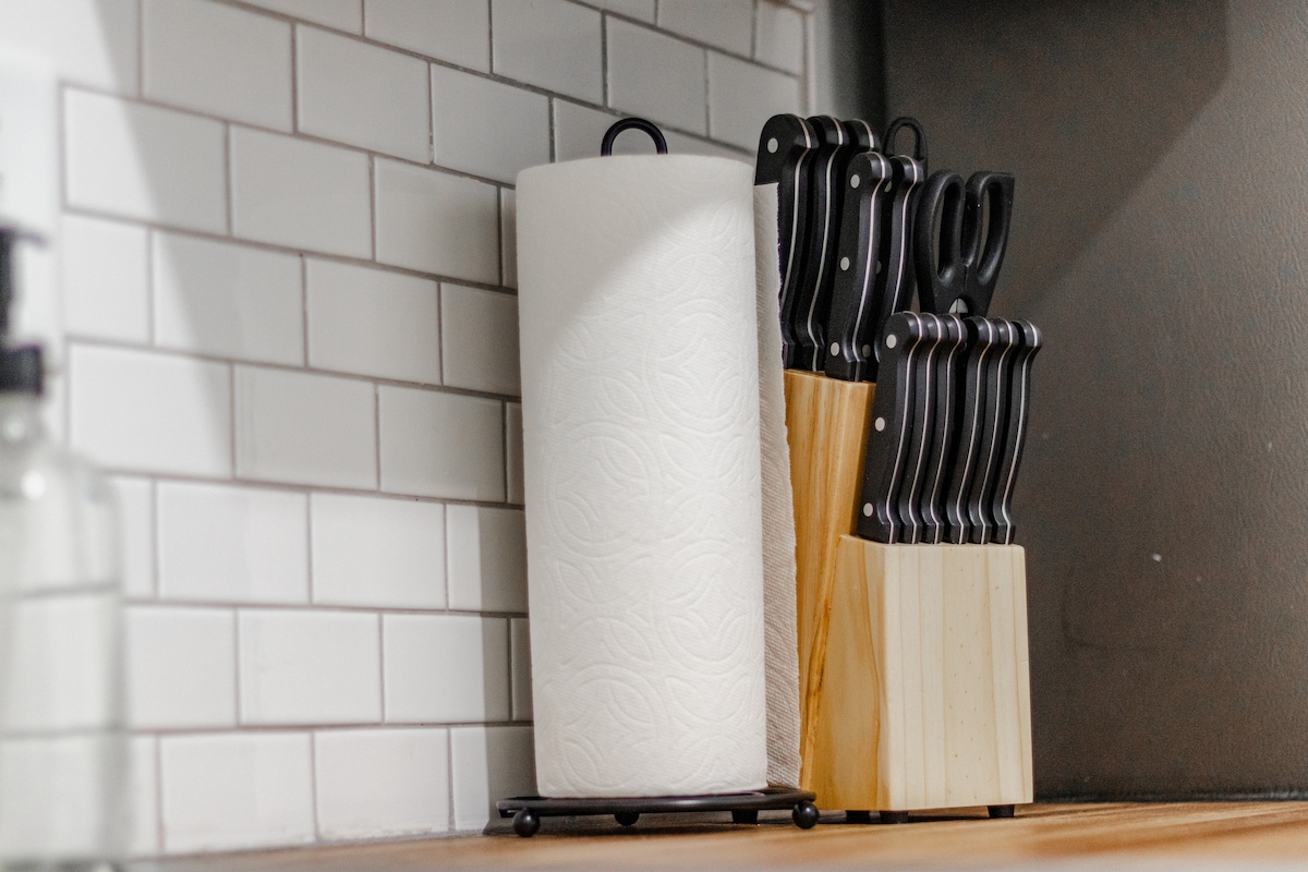A well-organized kitchen countertop showcases a roll of paper towels beside a set of black-handled kitchen knives stored in a wooden block. The backsplash features classic white subway tiles, enhancing the clean and functional kitchen space.