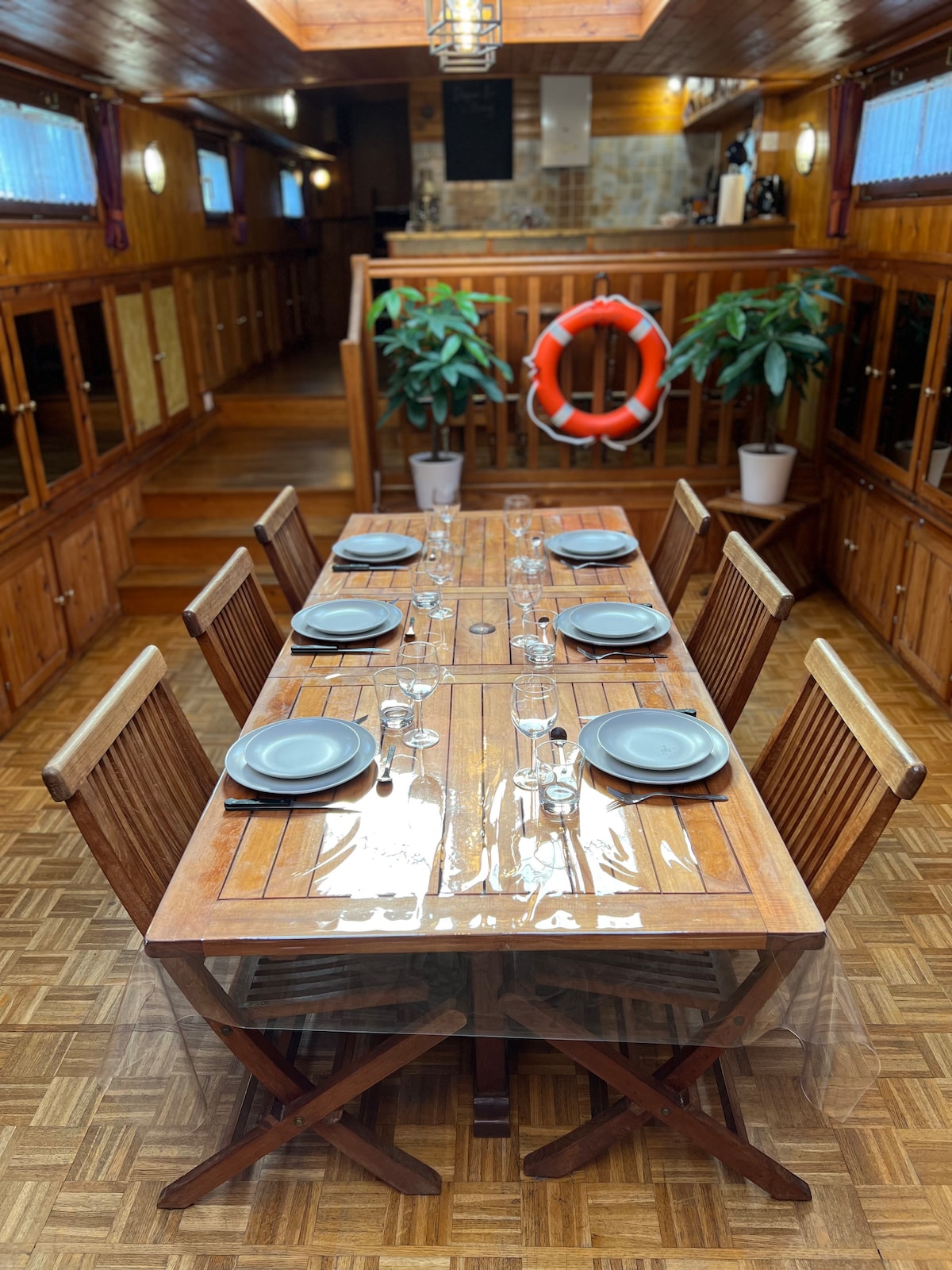 A wooden dining table is set for eight, featuring plates and wine glasses neatly arranged. Flanking the table, six chairs provide ample seating. The interior is illuminated by soft lighting, with potted plants and a life preserver decoratively positioned in the background.
