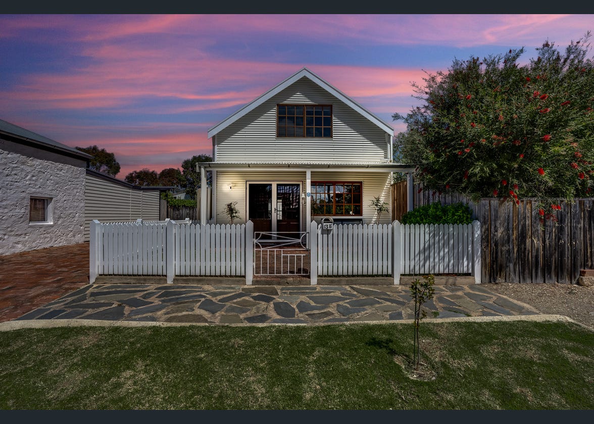 The exterior of a modern architectural home is presented, featuring a white picket fence and a stone pathway leading to the entrance. The façade has large windows that reflect the sunset colors, while the garden is well-maintained, providing a welcoming atmosphere.