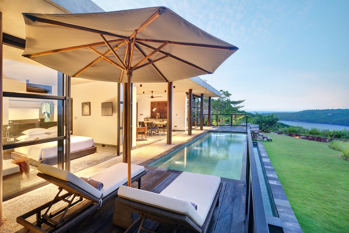 The outdoor space features a private infinity pool bordered by a sleek wooden deck. Two lounge chairs sit under a large umbrella, offering shade. The background showcases scenic views of Gamat Bay, while the villa's interior is visible through expansive glass doors.