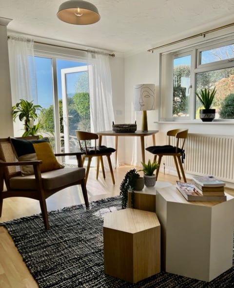A bright living space is equipped with a round wooden table surrounded by four mid-century style chairs. Sunlight filters through a glass door leading to the patio, with greenery visible outside. A textured rug adds warmth to the room, complemented by a stylish lamp and decorative plants.