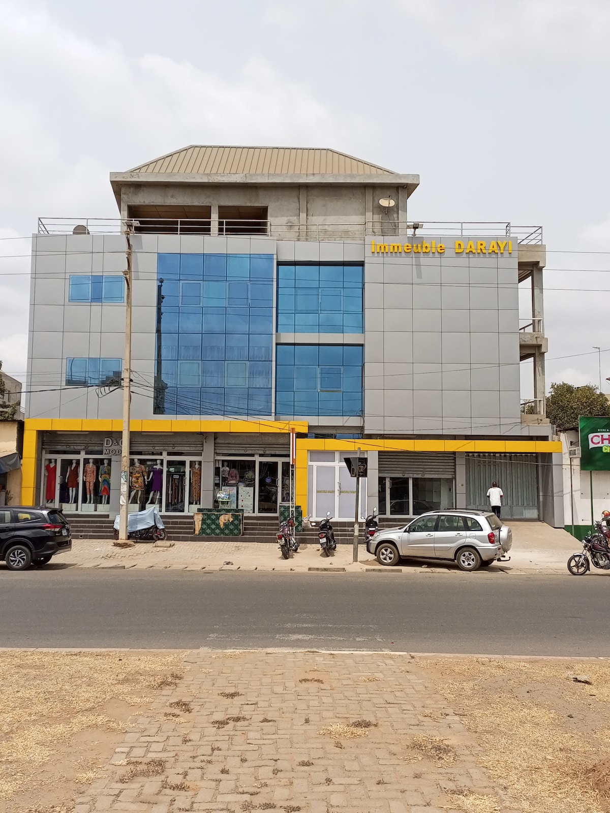 A modern multi-story building is observed, featuring a sleek glass facade with large, reflective windows. The entrance is marked by a bright yellow awning, indicating the business below. Surrounding elements include nearby shops and parked vehicles along the street.