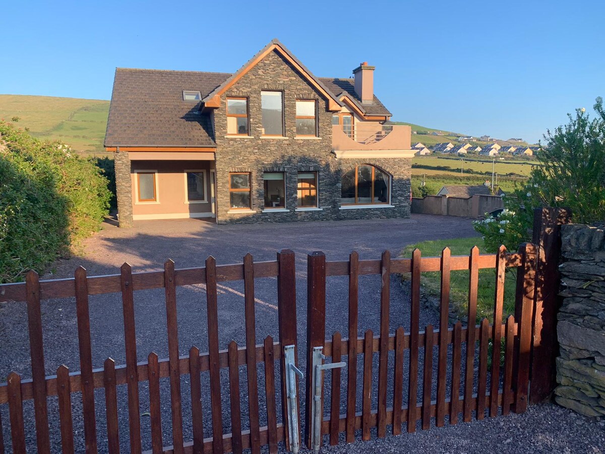 A modern two-story house is framed by landscaped greenery and a wooden picket gate. Large windows reflect natural light and showcase the surroundings. A gravel driveway provides access to the entrance, with scenic hills visible in the background.