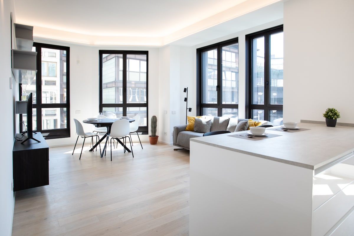 A bright living space is depicted, featuring a modern open-plan kitchen with a light countertop. A dining area with a round table and four chairs sits adjacent to a cozy seating area, highlighted by large windows that allow natural light to fill the room.