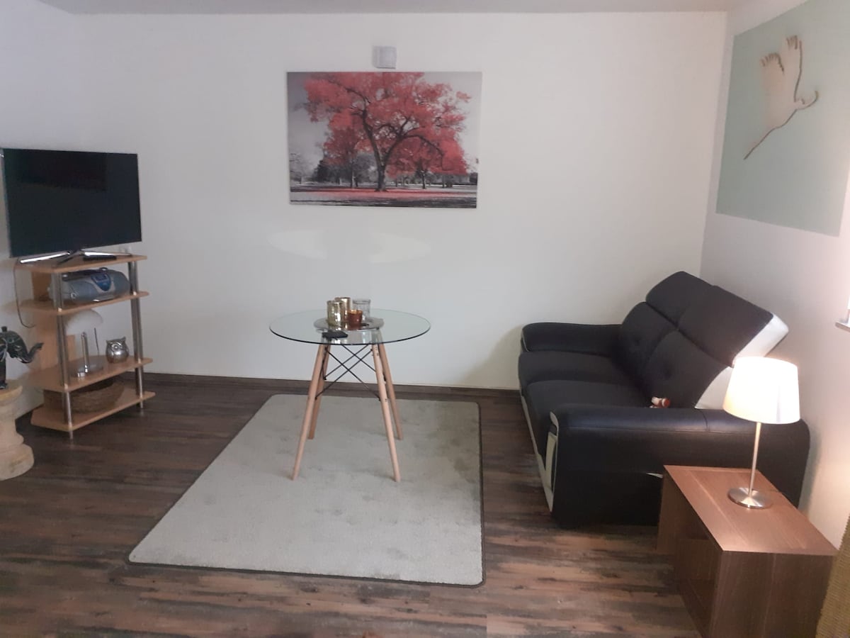 A living area is showcased with a modern black sofa positioned next to a glass-top table. A media unit with a television is located nearby, along with a softly lit floor lamp. The wall is decorated with artwork featuring a red tree, enhancing the space's character.