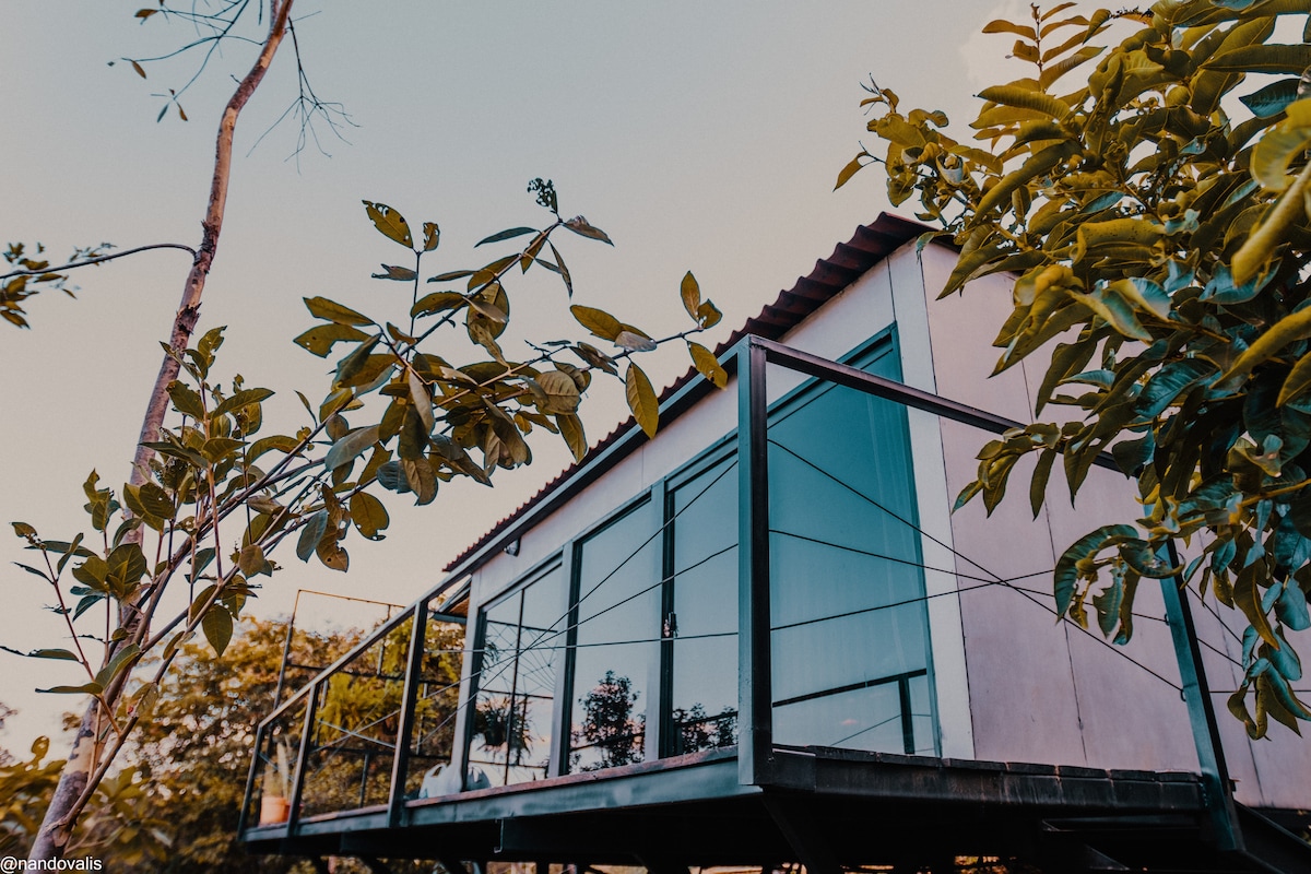 A tiny house is depicted, elevated on a metal structure, surrounded by lush greenery. The exterior features large glass doors and a wooden roof. A small balcony with a glass railing overlooks the natural landscape, emphasizing a connection to the environment.