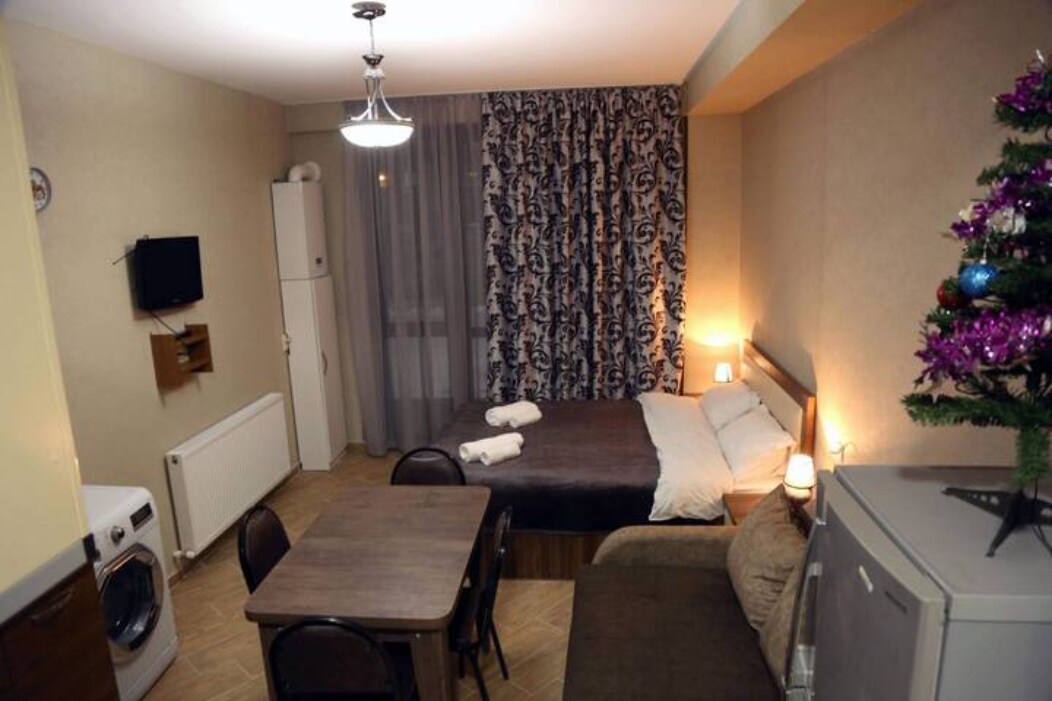 A well-equipped studio apartment features a spacious bed dressed in neutral tones, complemented by a decorative wall with patterned curtains. A dining table with chairs is positioned near a fridge and washing machine, creating a functional living space. Soft lighting enhances the overall ambiance.