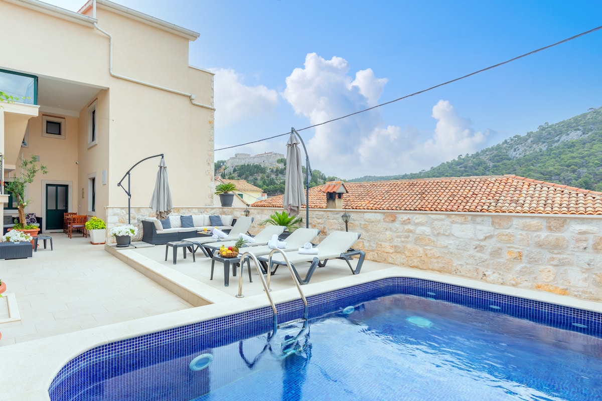 A modern outdoor area features a rectangular pool surrounded by lounge chairs and umbrellas. Lush greenery is seen in planters, with terracotta-tiled rooftops and mountains in the backdrop. Bright clouds fill the sky, creating an open and airy atmosphere.