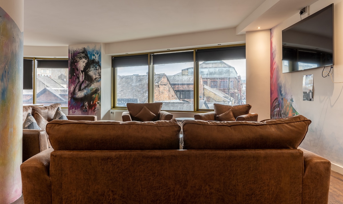 A spacious living area features a modern arrangement of comfortable brown sofas facing a wall-mounted television. Large windows provide ample natural light, revealing views of the cityscape. Colorful artwork adorns the walls, adding a vibrant touch to the contemporary design.