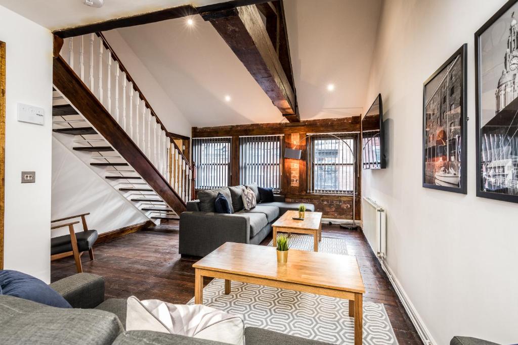 A spacious lounge area features a comfortable grey sofa and two wooden coffee tables positioned on a patterned rug. Large windows allow natural light to fill the room, while exposed wooden beams add an architectural element. A staircase leads to the upper level, complementing the rustic charm.