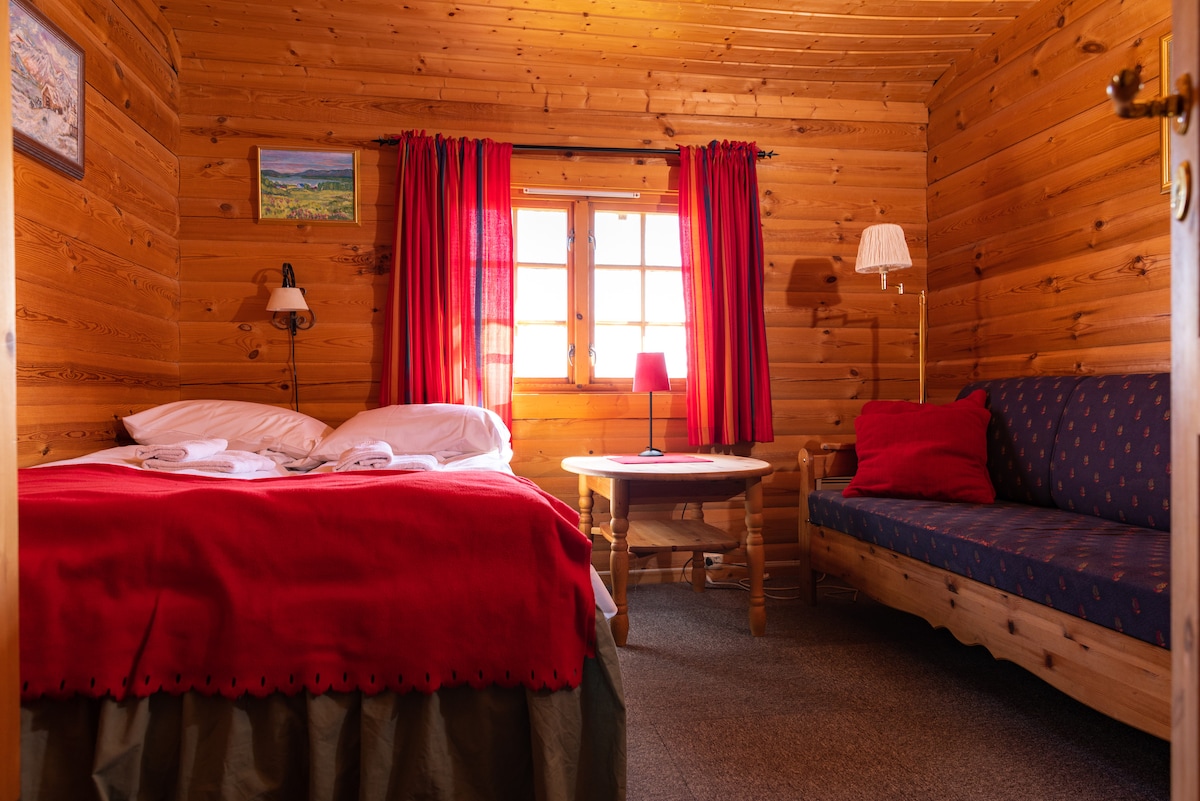 A cozy room features warm wooden paneling and bright red curtains framing a window. A spacious bed with white linens is complemented by a small round table with a lamp. A comfortable seating area with a sofa and a decorative pillow is also present.